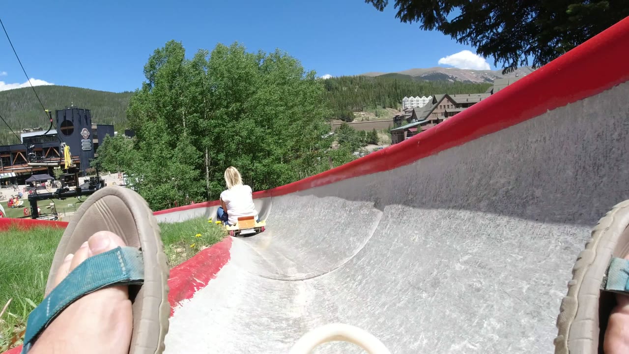 Winter Park, Colorado slide track view with the rider's sandal close to camera, the child farther ahead, a red track rim, leafy trees, and resort buildings under bright daylight.