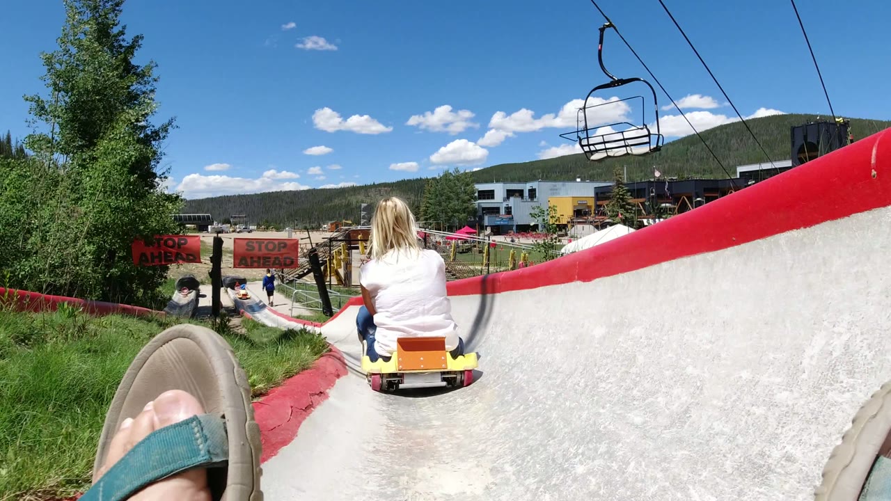 At 0:01 in Winter Park, Colorado, the yellow cart ahead moves closer to red stop ahead signs while the concrete channel, trees, fair buildings, and mountain horizon remain visible.