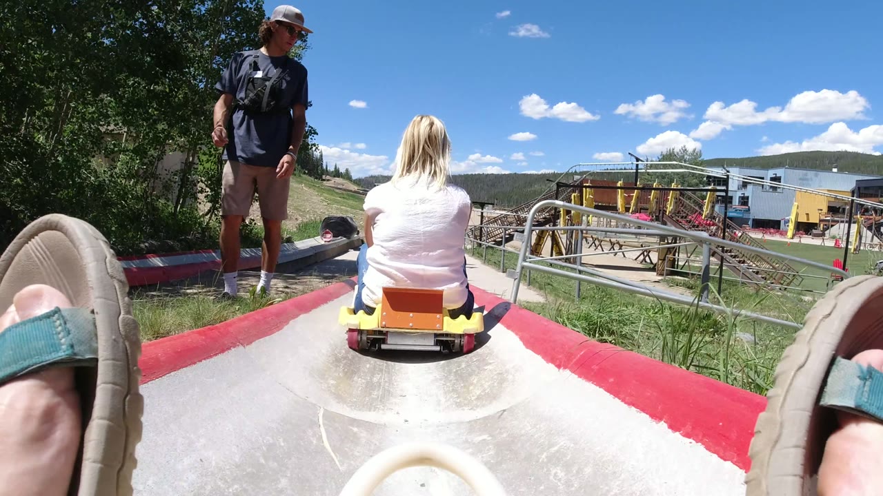 Winter Park, Colorado slide scene with a woman in a white shirt riding away from the camera, a standing man to the left, foreground sandals, and hillside railings beyond the chute.