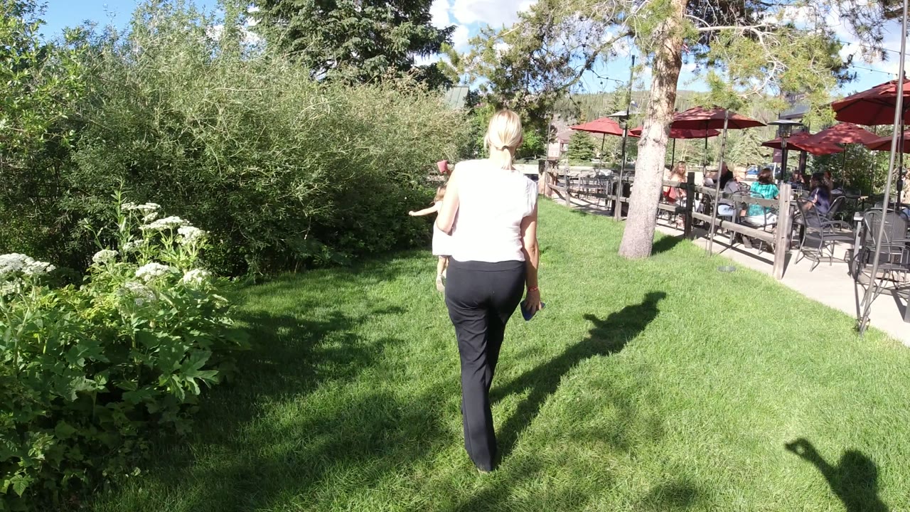 In Winter Park, Colorado, the woman moves farther into the lawn as the child is partly hidden by her position, with patio tables and red umbrellas visible beyond the trees.