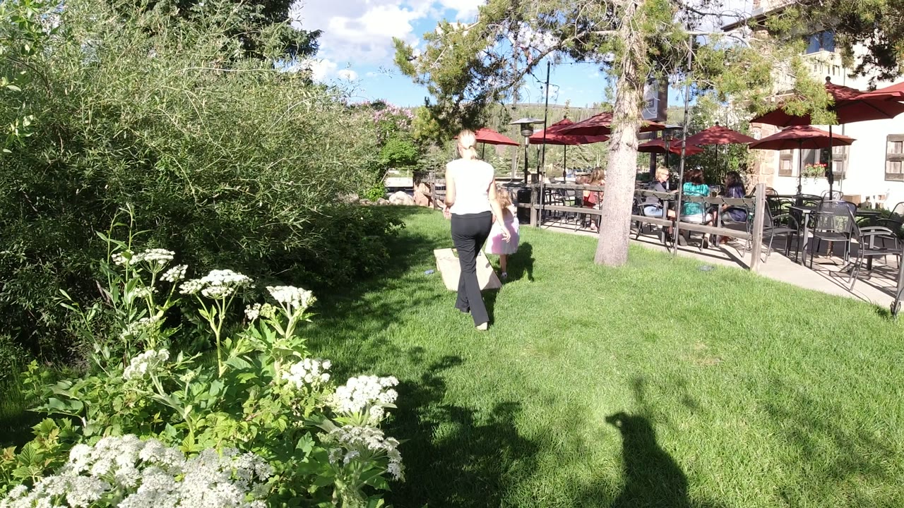 In Winter Park, Colorado, the woman moves closer to the child and white bean bag toss board, with bright grass, shrubs, trees, and red patio umbrellas behind them.