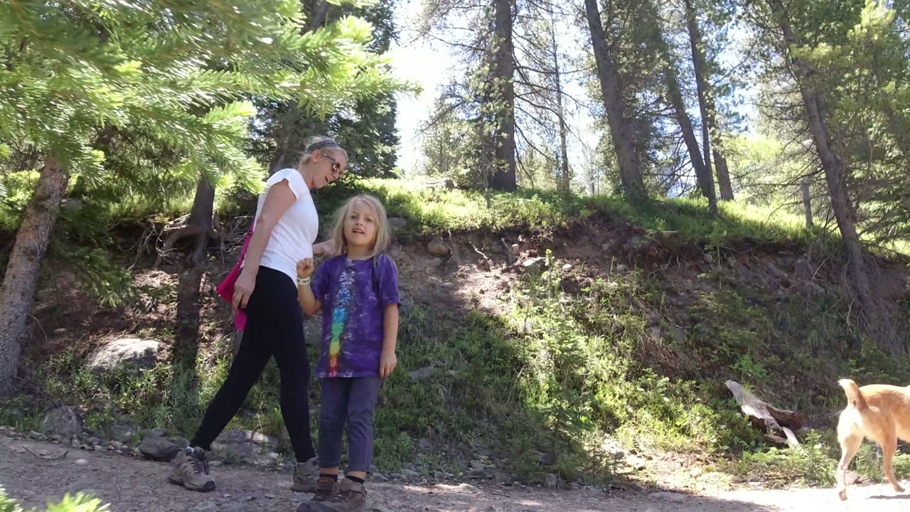 A woman in a white shirt and a blonde child in a purple shirt stand on a sunlit dirt trail in Winter Park, Colorado, with pine trees and rocks behind them.