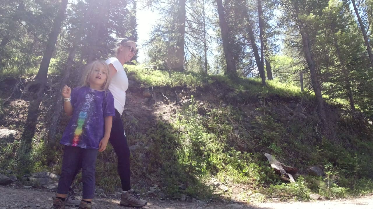 The child faces the camera near the center of the Winter Park trail while the woman stands behind and a tan dog moves through the sunlit path.