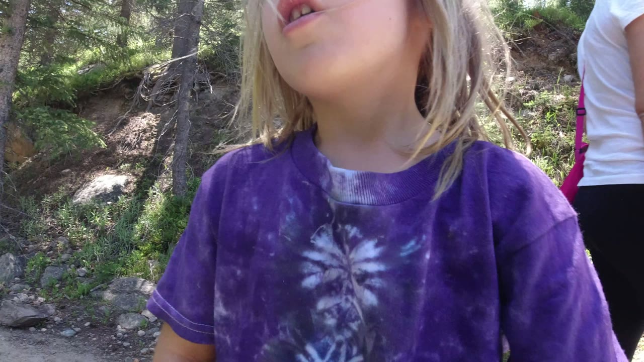 A blonde child in a purple tie-dye shirt fills the foreground on a wooded trail in Winter Park, Colorado, with part of an adult walking behind her.