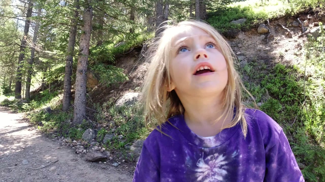The child looks upward while standing or walking on a sunlit dirt path in Winter Park, Colorado, with pine trees and a rocky slope behind her.