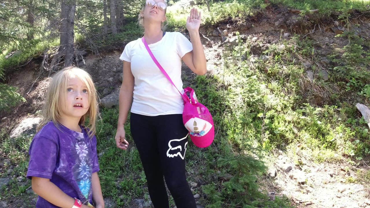 A blonde child in a purple shirt stands in front of an adult woman with a pink bag on a sunlit wooded slope in Winter Park, Colorado.