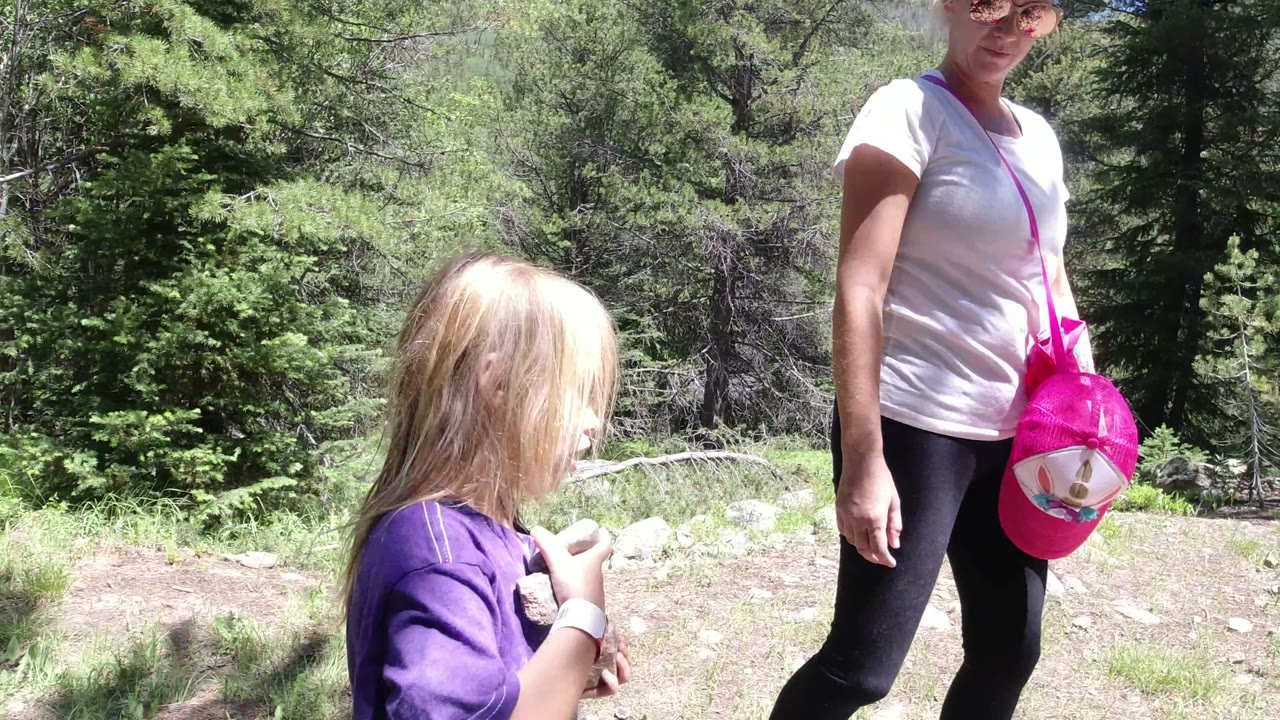 In Winter Park, Colorado, a blonde child in a purple shirt holds a small object while an adult with a bright pink bag stands nearby on a sunny forest trail.
