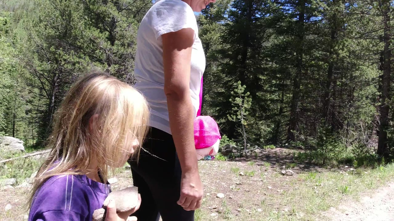 A close view in Winter Park, Colorado shows the child walking with hair across the face while the adult passes beside them against a green pine forest.
