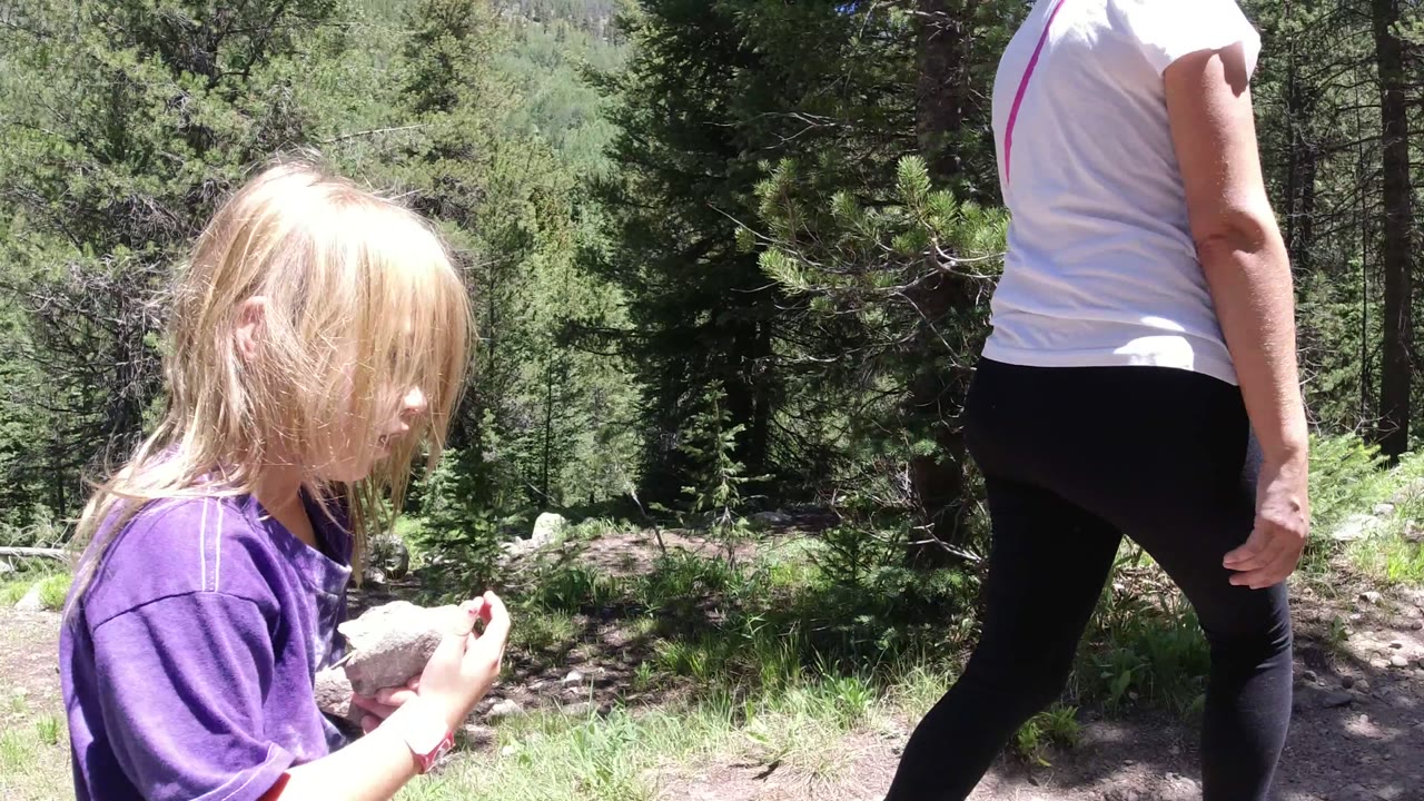 An adult in a white shirt and black leggings walks along a dirt path in Winter Park, Colorado, with the child partly visible at the left edge and forest in the background.