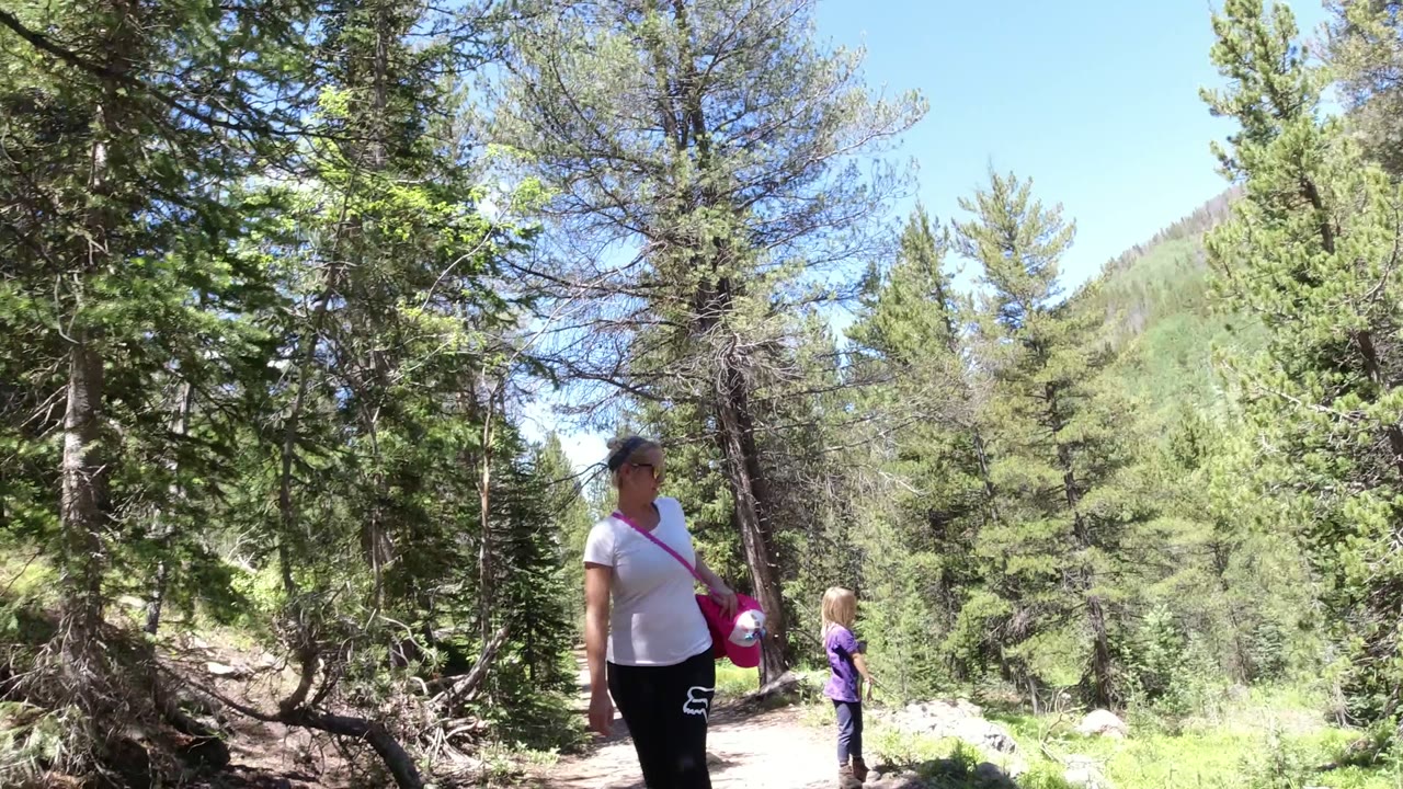 The adult stands near the trail edge while the child is farther down the path, surrounded by conifer trees and bright green forest in Winter Park, Colorado.