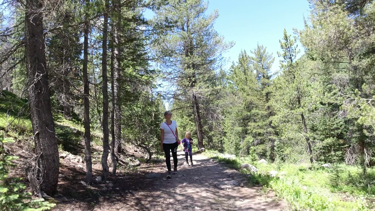Two tourists walk along a shaded dirt trail in Winter Park, Colorado, with tall trees on both sides and bright green vegetation beyond the path.