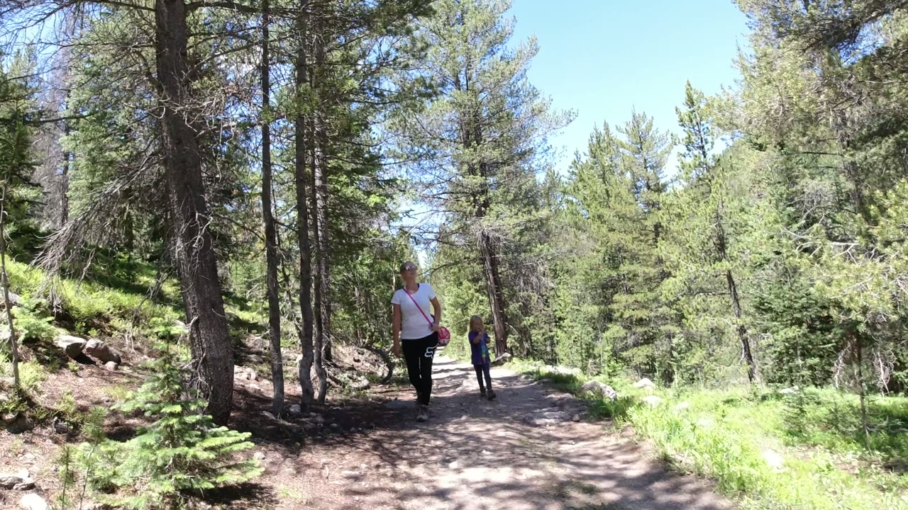 A woman in a white top walks closer to camera on a Winter Park, Colorado forest trail while a second person follows farther back under tall trees.