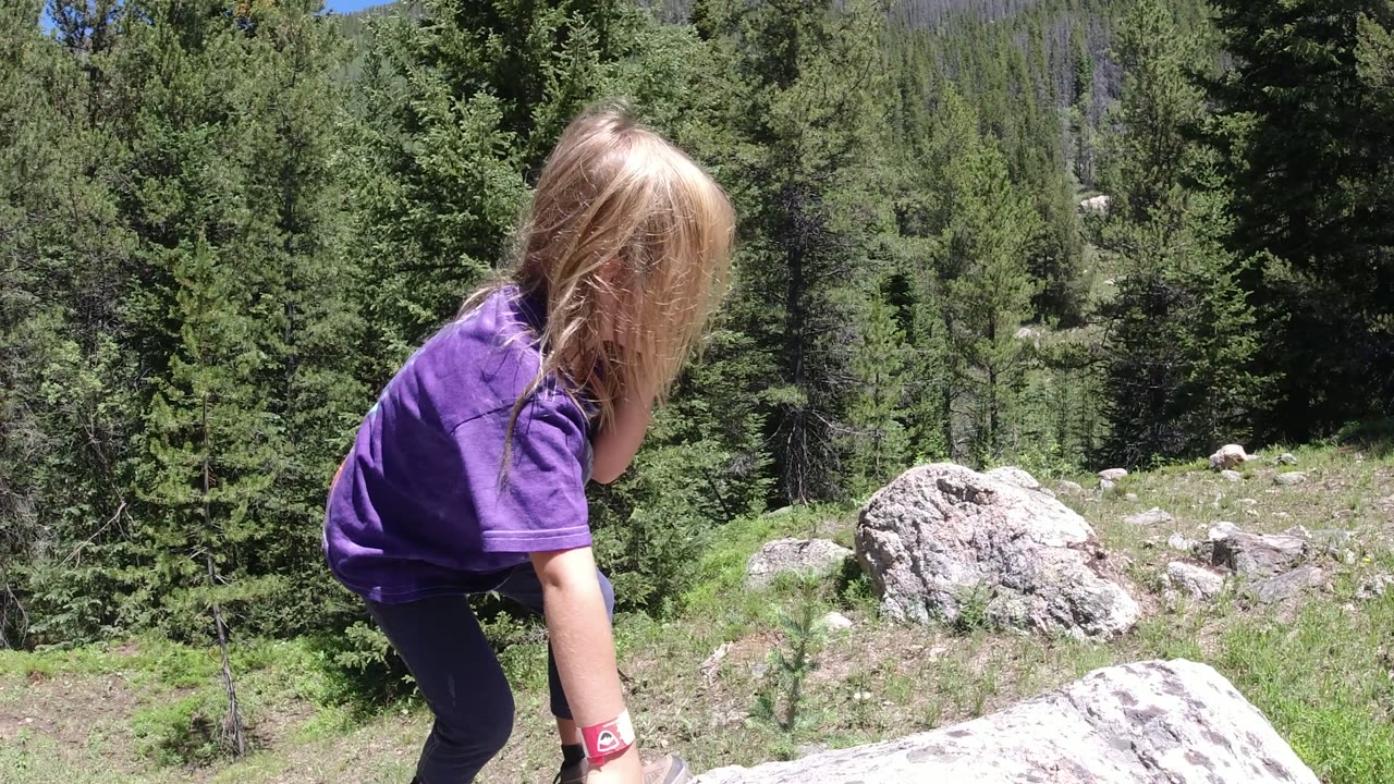 A blond child in a purple shirt leans forward beside a pale rock in Winter Park, Colorado, with dense green trees behind them.