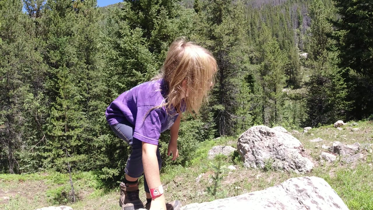 The child bends lower toward the foreground boulder in Winter Park, Colorado, with conifer trees and gray rocks across the hillside.