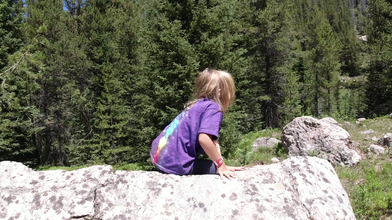 At 0:00, a blond child in a purple shirt sits low beside a large pale boulder in Winter Park, Colorado, with evergreen trees filling the background.