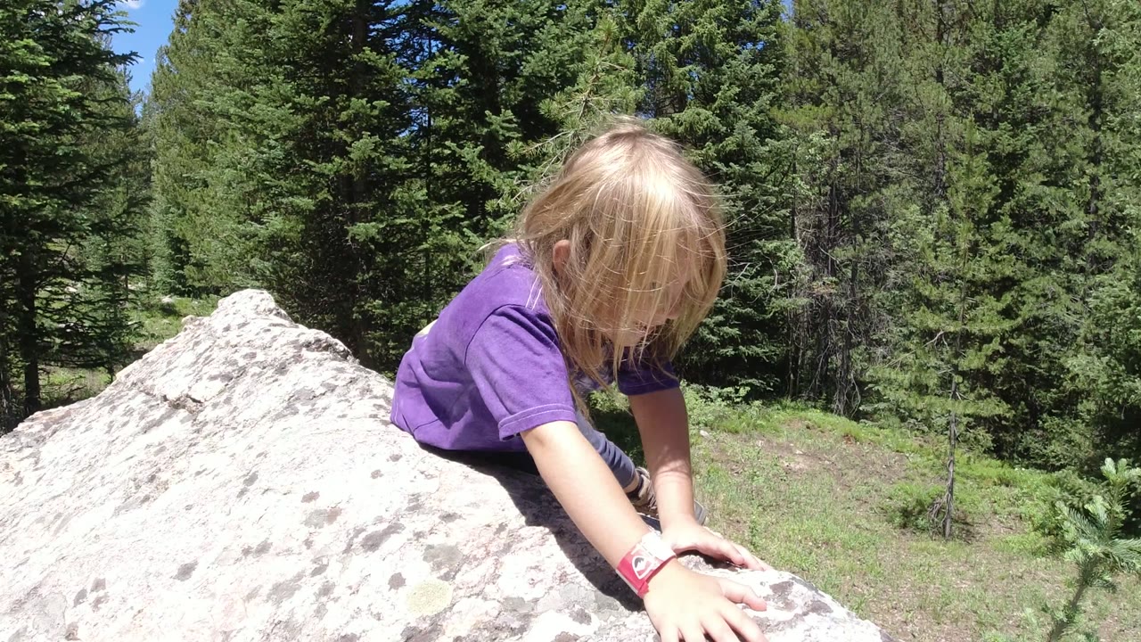 At Winter Park, Colorado, a young blond girl in a purple shirt reaches down across a large white rock with pine trees behind her.