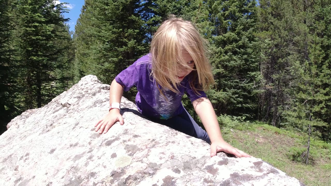 At Winter Park, Colorado, the girl braces both hands on the pale rock while her hair falls forward and evergreen trees fill the background.
