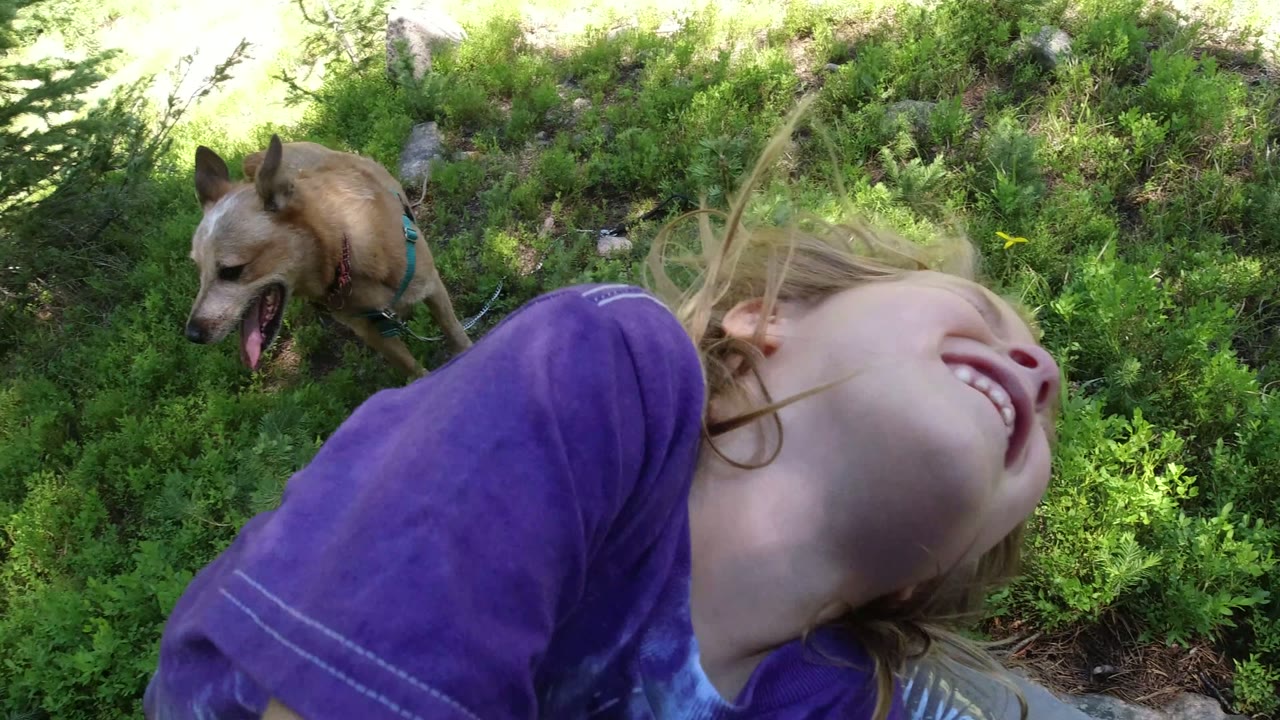 Blonde girl laughing near a tan dog in green woods.