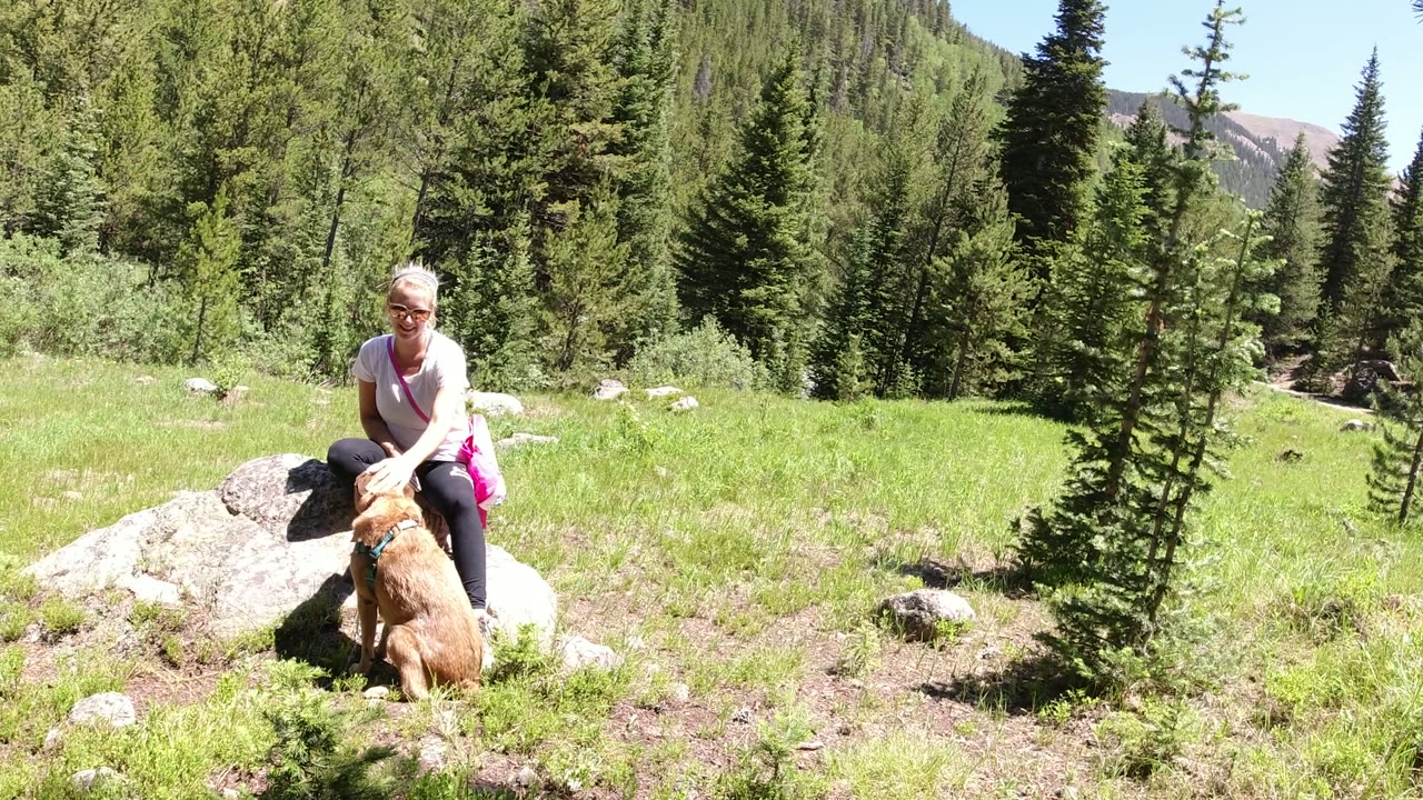 Two seconds in, the woman remains by the boulders with the tan dog in front of her, while a small evergreen stands near center frame and forested slopes fill the background in Winter Park, Colorado.