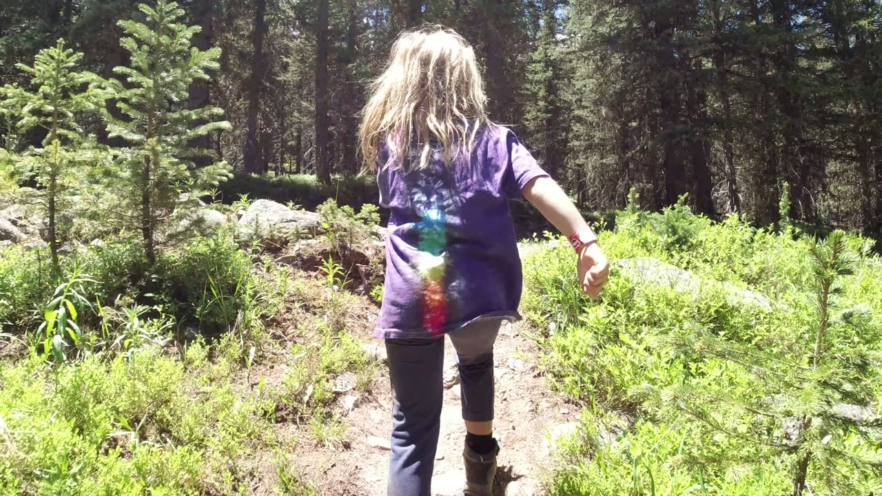 The girl continues along the Winter Park trail, centered from behind with bright green undergrowth and dark trees beyond.