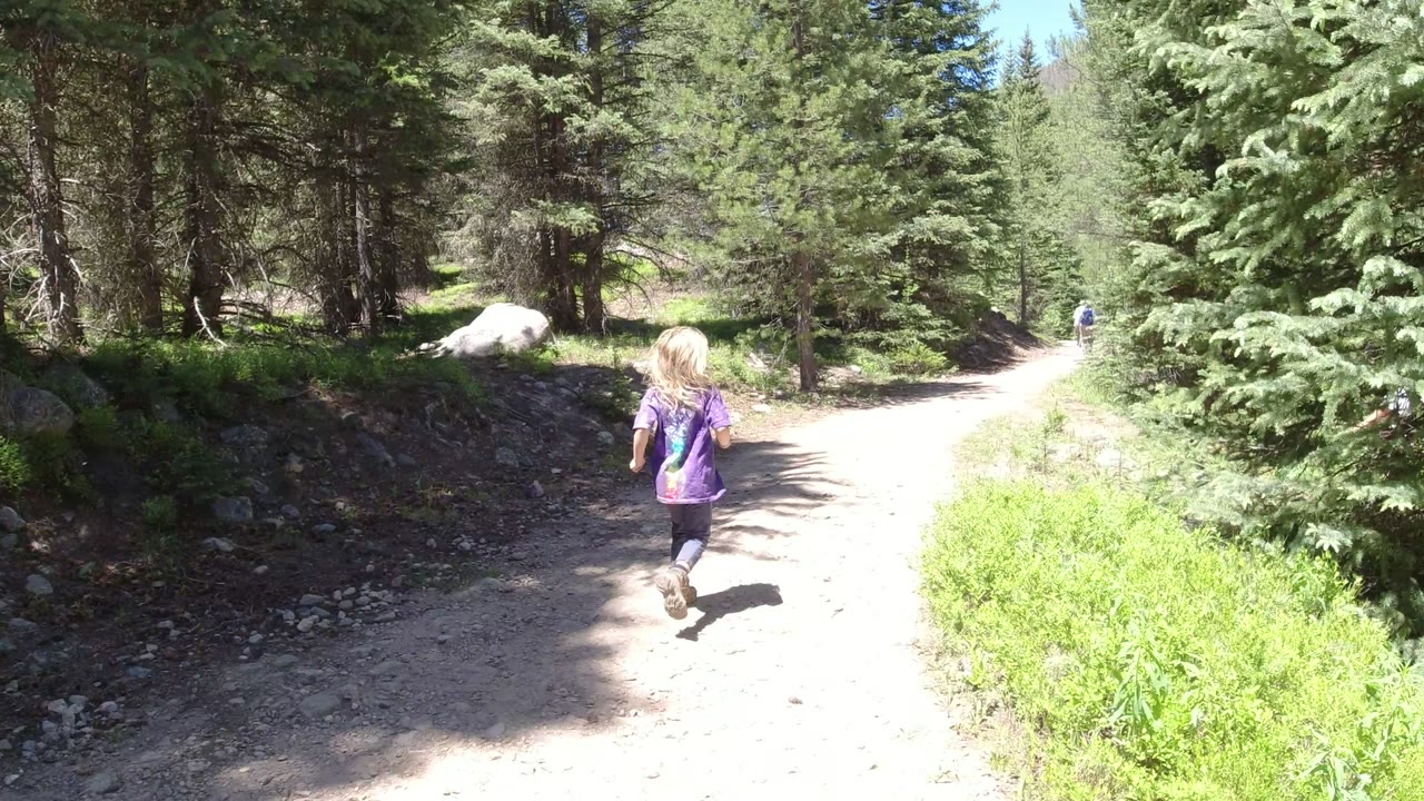 The child continues up the bright dirt trail, with dense evergreen trees and a large pale rock visible beside the path in Winter Park, Colorado.