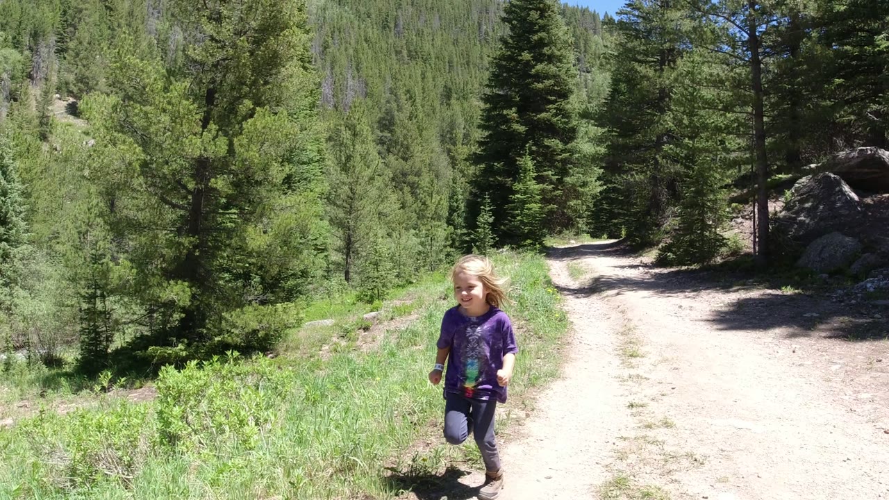 The child is closer in the frame on the dirt path in Winter Park, Colorado, with conifer trees, grass, and boulders behind and beside the road.
