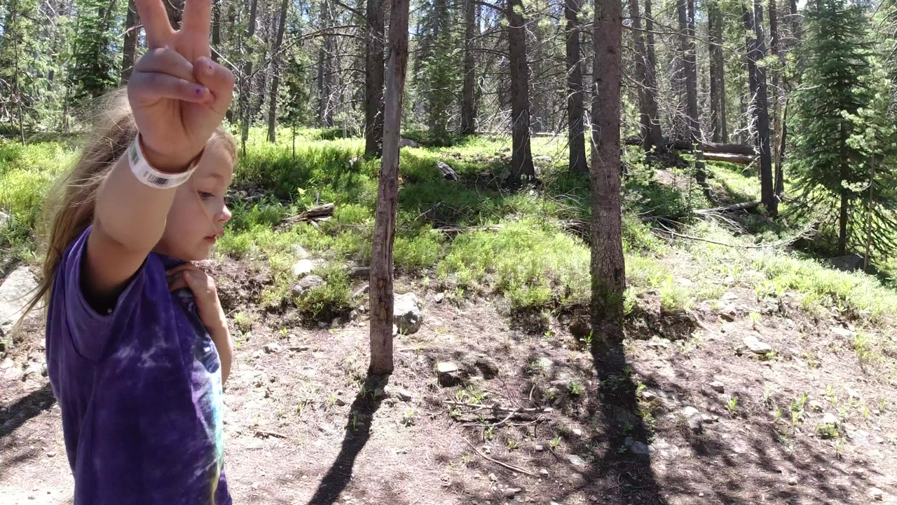 At the start of the Winter Park, Colorado clip, a young child in a purple shirt is cropped at the left edge while holding up two fingers near a dirt path and tall trees.
