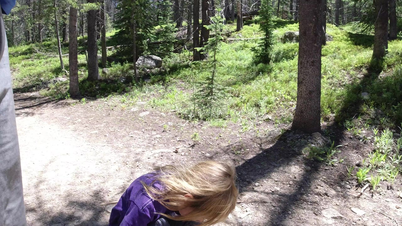 At 0:01 in Winter Park, Colorado, the child remains low in the foreground while a sunlit dirt trail, tree trunks, shrubs, and a gray pant leg at the right edge fill the frame.