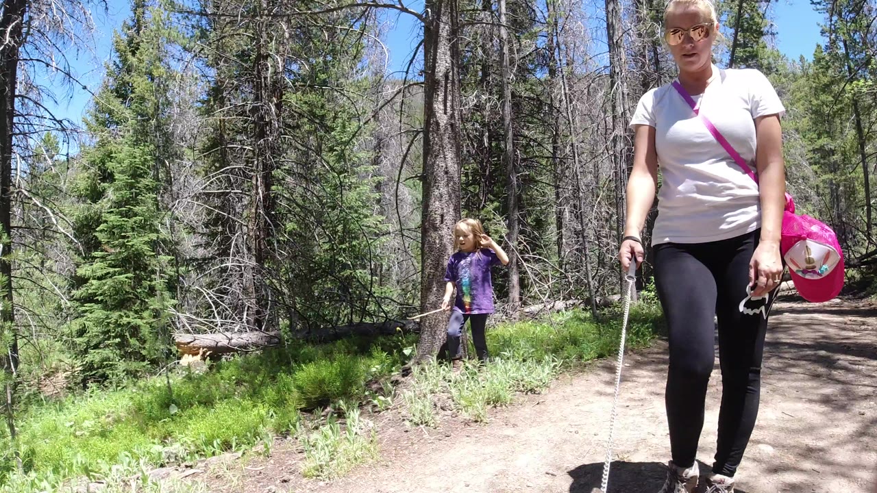 In Winter Park, Colorado, a woman walks close to the camera on a dirt forest trail while a young girl stands farther back near green grass and tall trees.