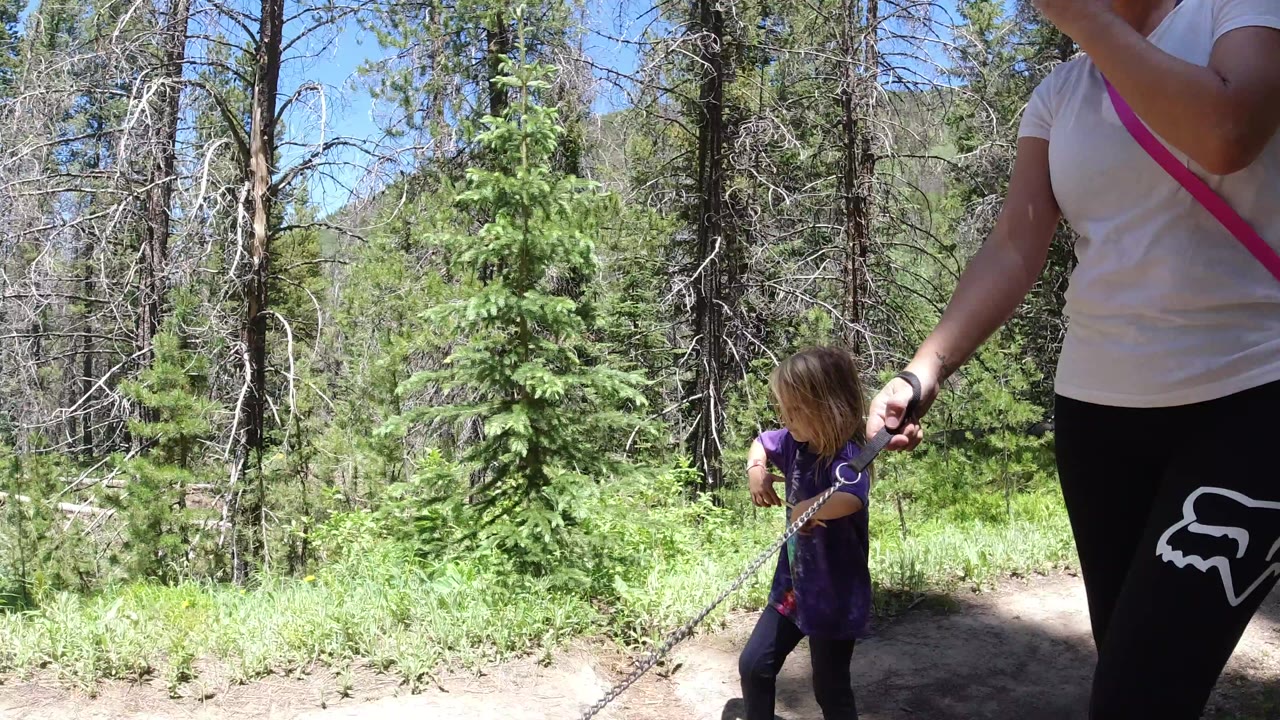 A child in a purple shirt walks near the edge of a dirt path while the adult woman's torso and hand holding a chain or rope fill the right foreground in Winter Park, Colorado.