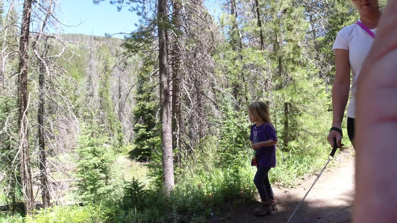 In Winter Park, Colorado, the child walks along the dirt path in front of evergreen trees while the adult remains partly cropped at the right edge.