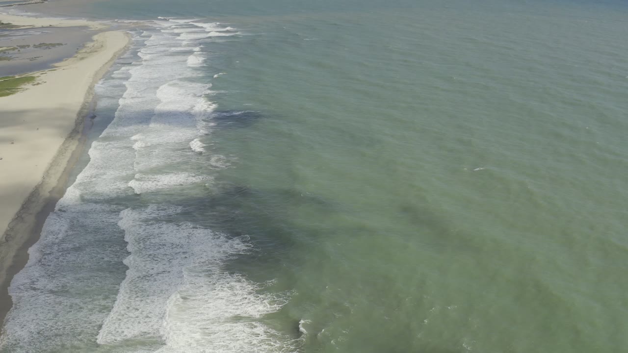 Aerial view over a wide Los Cabos beach with green-blue water, white surf bands, pale sand, and no visible people.
