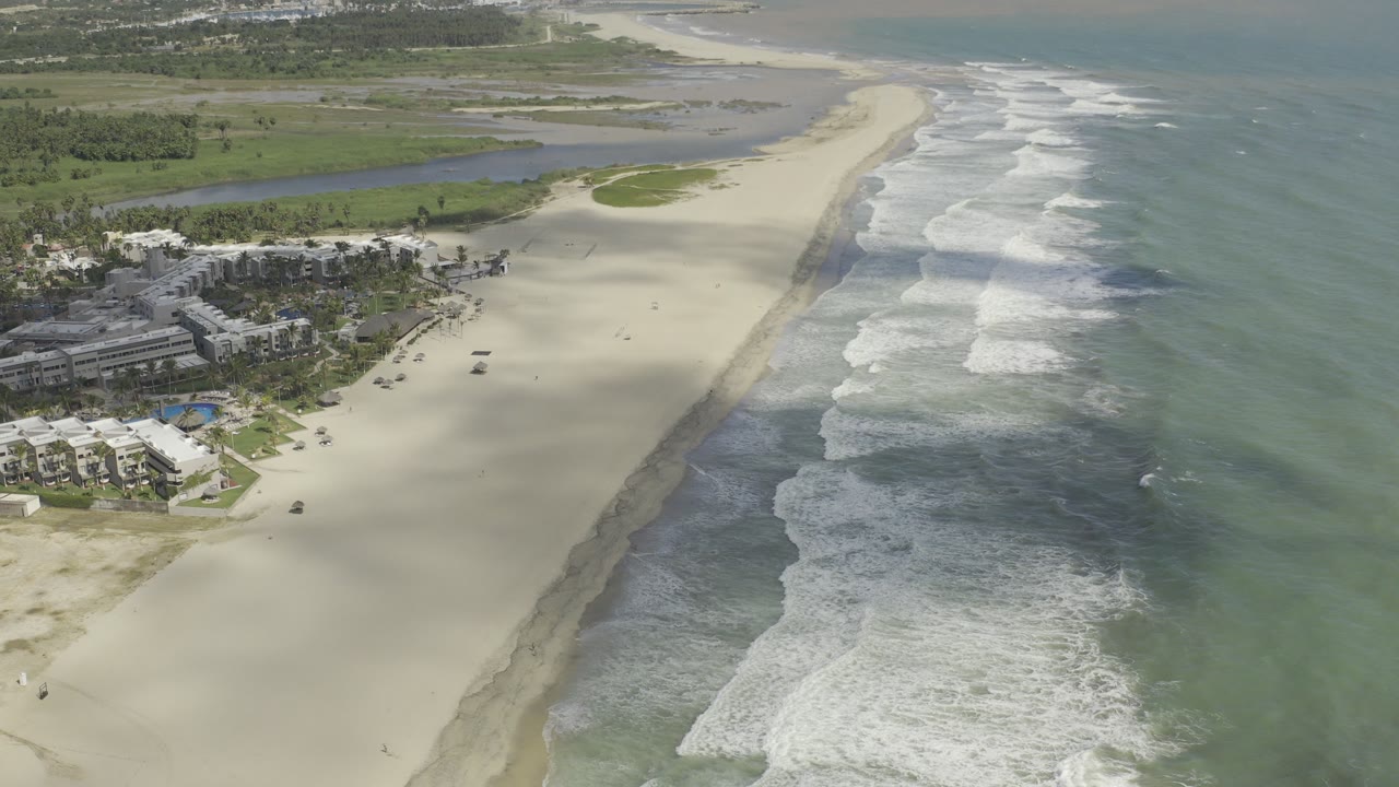 Aerial view over a Los Cabos beach with resort buildings at lower left, pale sand, parallel surf lines, lagoon water inland, and no visible people.