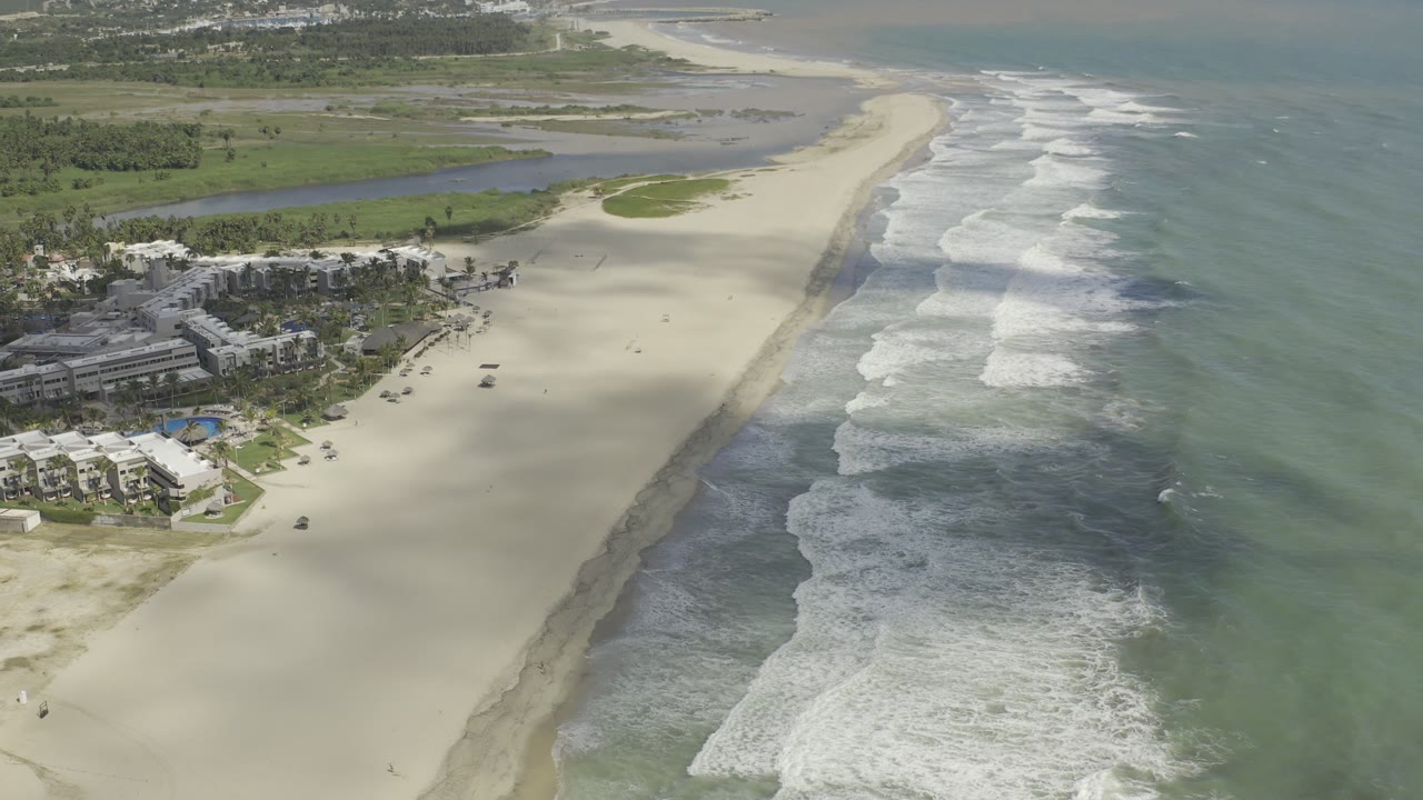 Aerial Los Cabos coastline frame with the resort buildings more centered, surf breaking along the right side, inland lagoon water behind the sand, and no visible people.