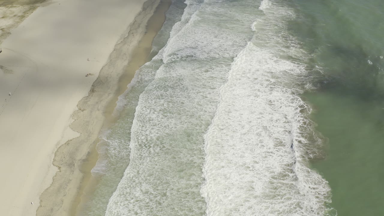 Aerial Los Cabos shoreline frame with a thicker white wave band spreading across wet sand and green water, with no visible people in this frame.