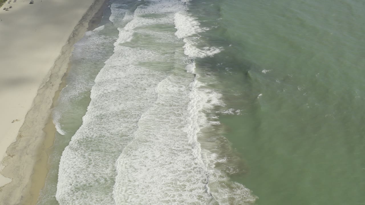 Overhead view of Los Cabos surf breaking in white bands beside pale sand and green water, with no visible people.