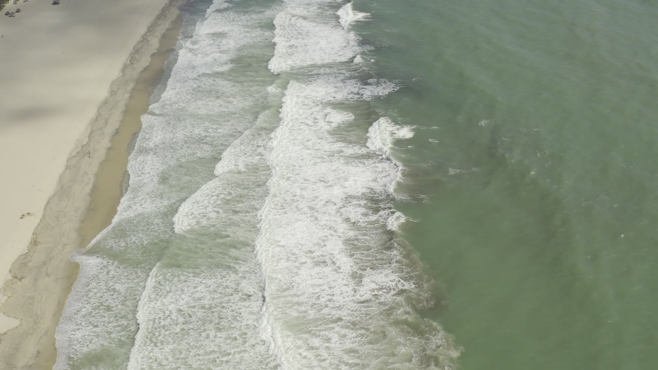 Aerial view of white surf breaking along a sandy beach beside green ocean water.