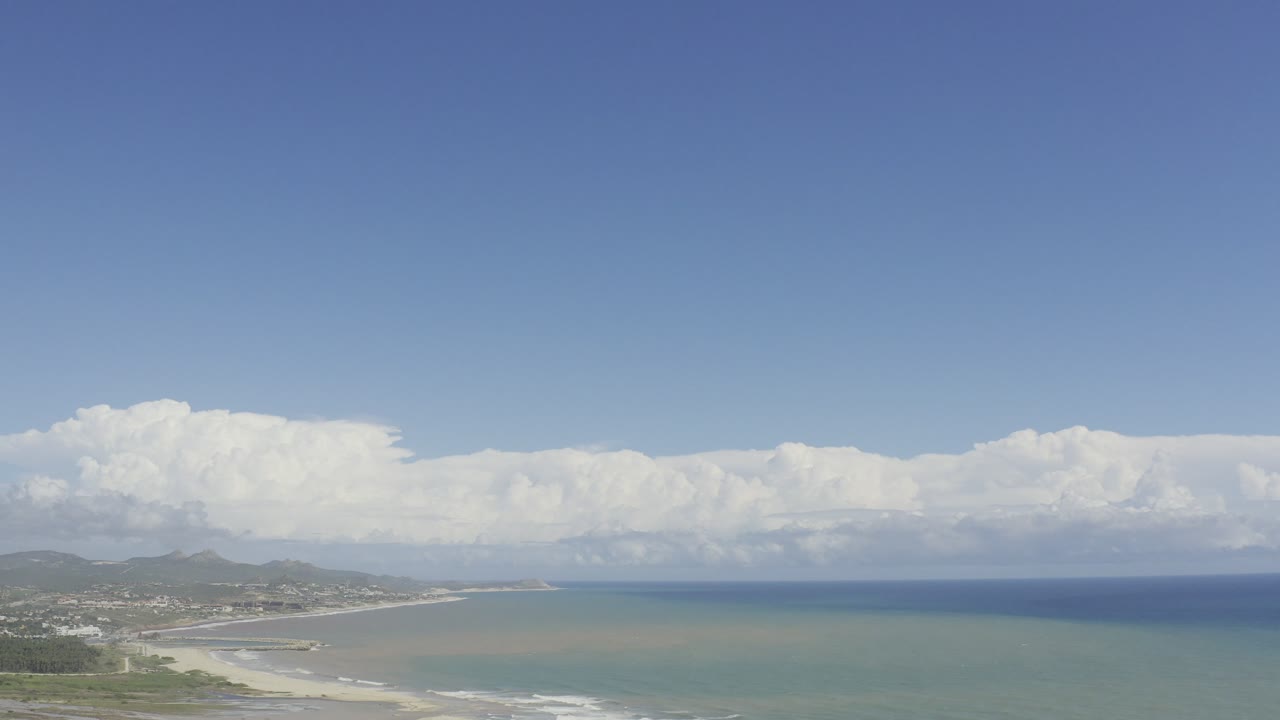 Aerial view in Los Cabos with the shoreline lower in frame, pale sand, thin white surf lines, distant hills, clouds, and no visible people.