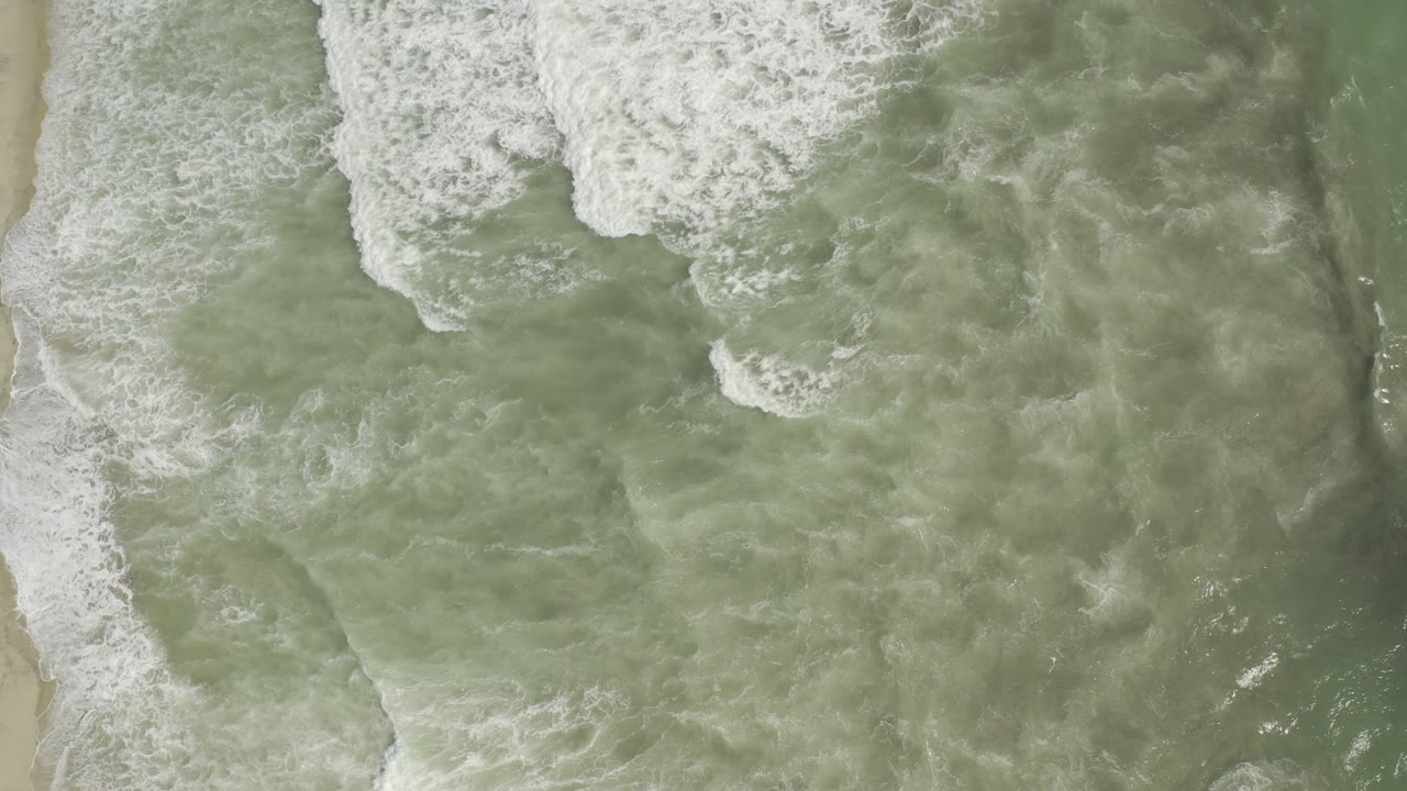 Top-down view of pale green surf and white foam reaching the sand in Los Cabos, Mexico, with no visible people.