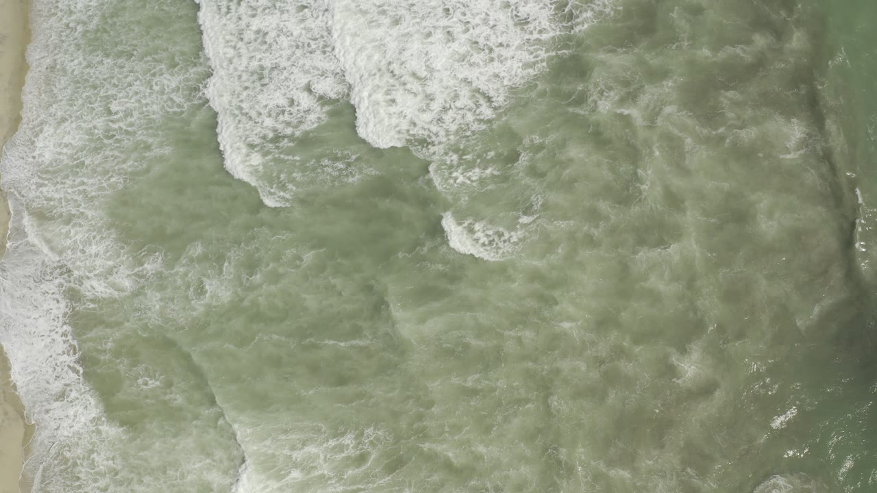 Aerial straight-down frame of white breakers spreading across muted green water near the Los Cabos beach, with no visible people.