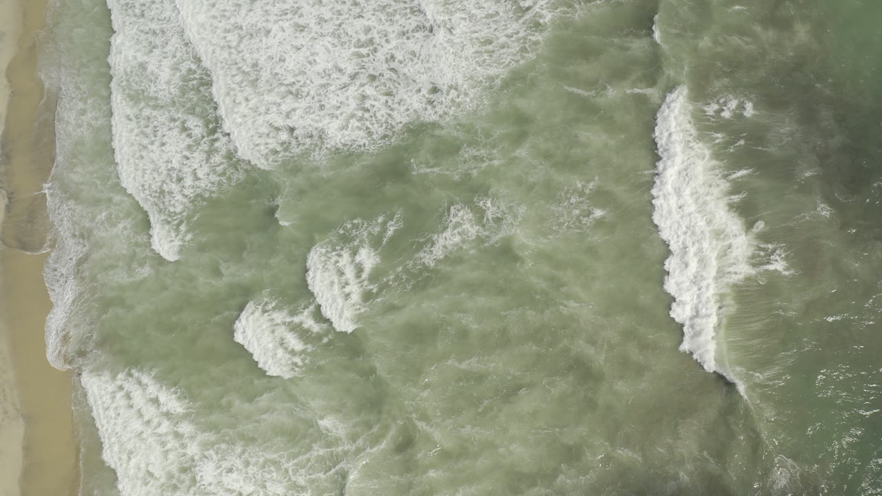 Overhead view of green waves and white foam reaching a tan Los Cabos beach, with no visible people.