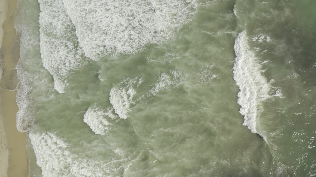 Aerial frame of white surf bands spreading across green water near the Los Cabos shoreline, with no visible people.