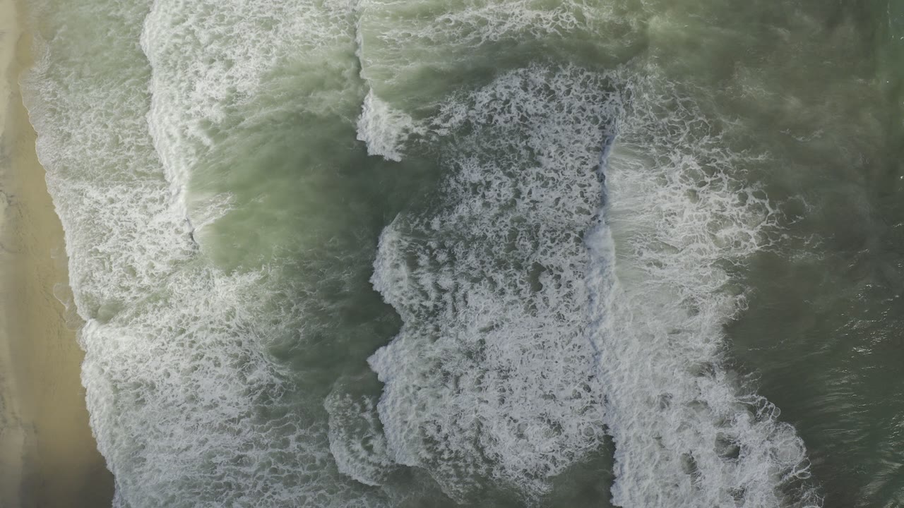 Overhead view of layered white surf and green-gray water beside pale sand in Los Cabos, Mexico, with no visible people.