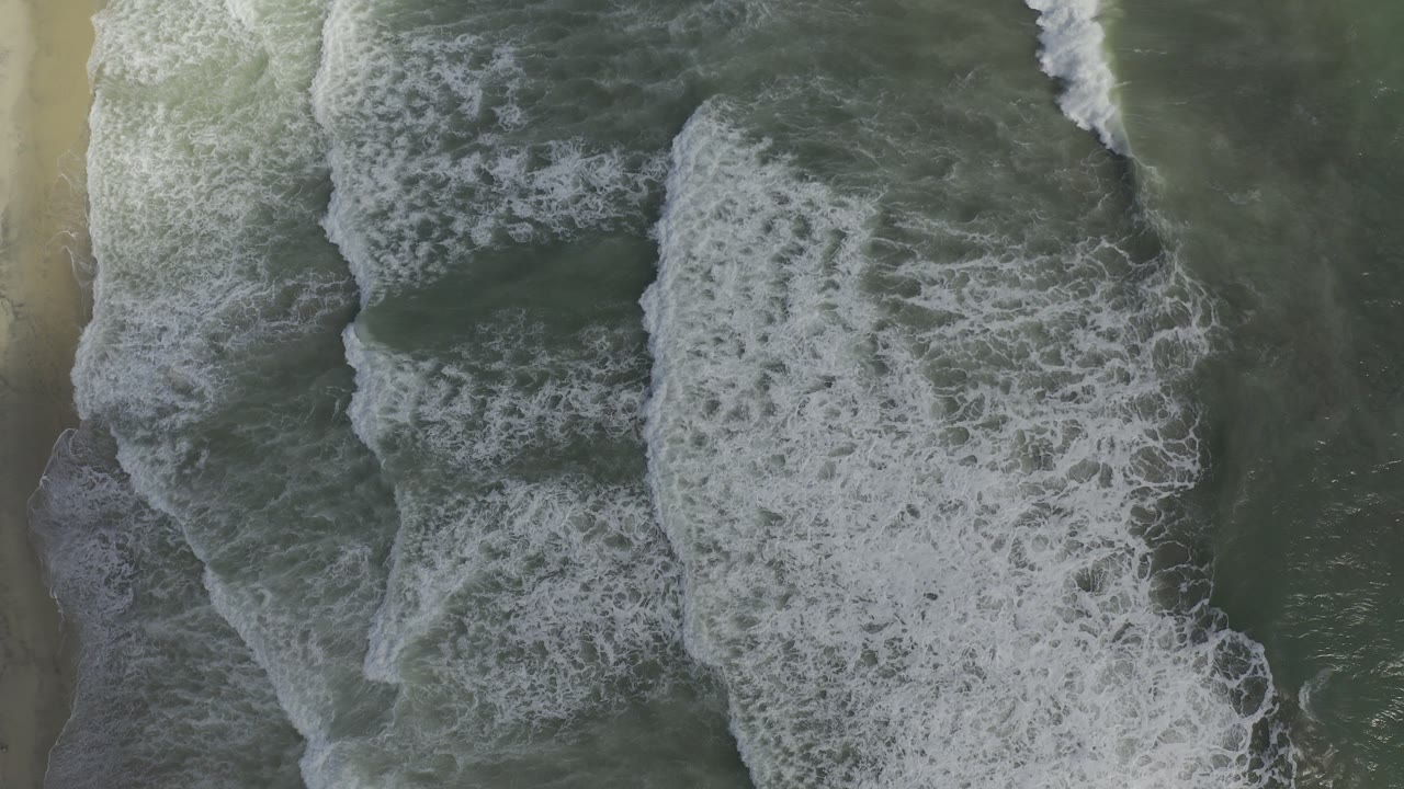 Overhead view of Los Cabos surf with white foam spreading across green water beside a pale sand edge, no visible people.