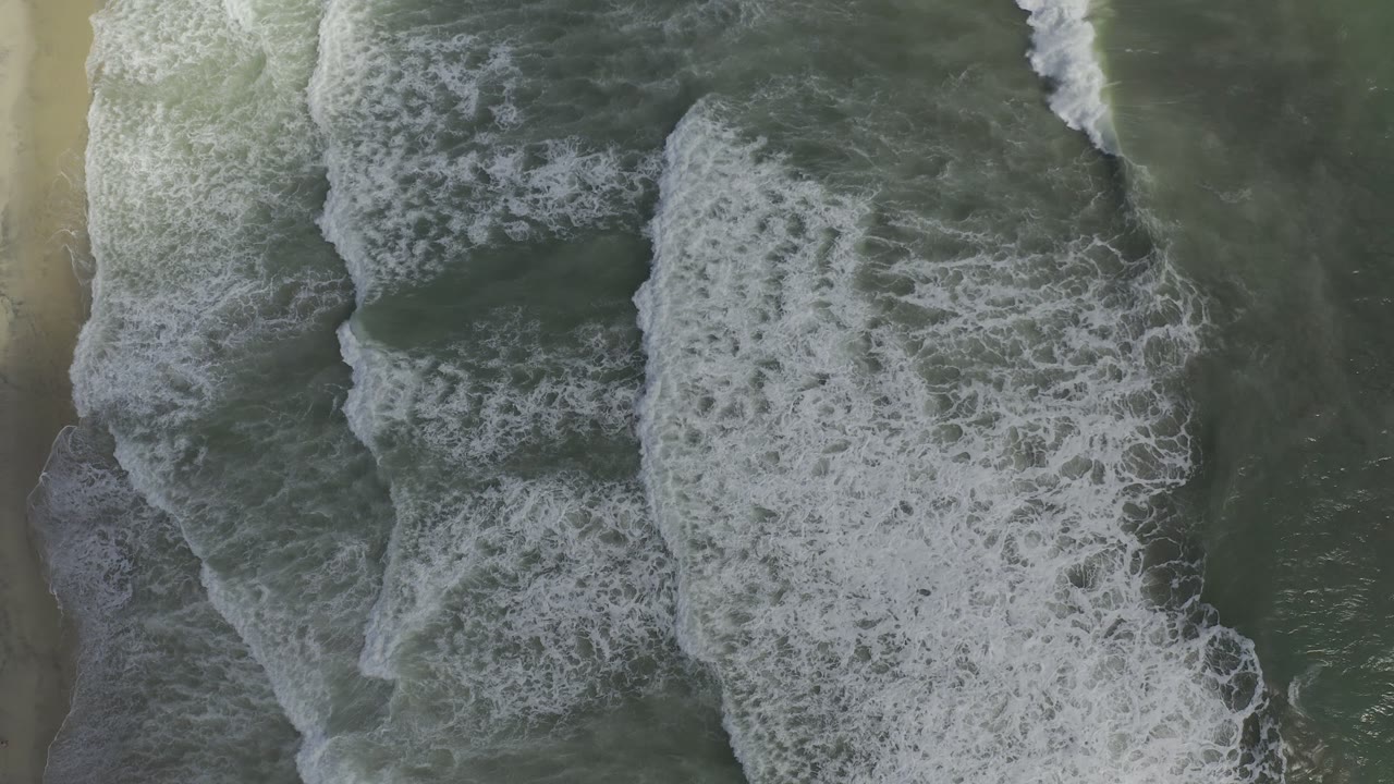 Aerial Los Cabos shoreline frame with a wide white breaker crossing green water and pale sand running along the side, no visible people.