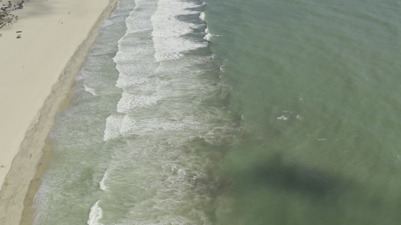 Aerial view over Los Cabos, Mexico with pale sand at left, green water at right, diagonal surf lines, and no visible people.