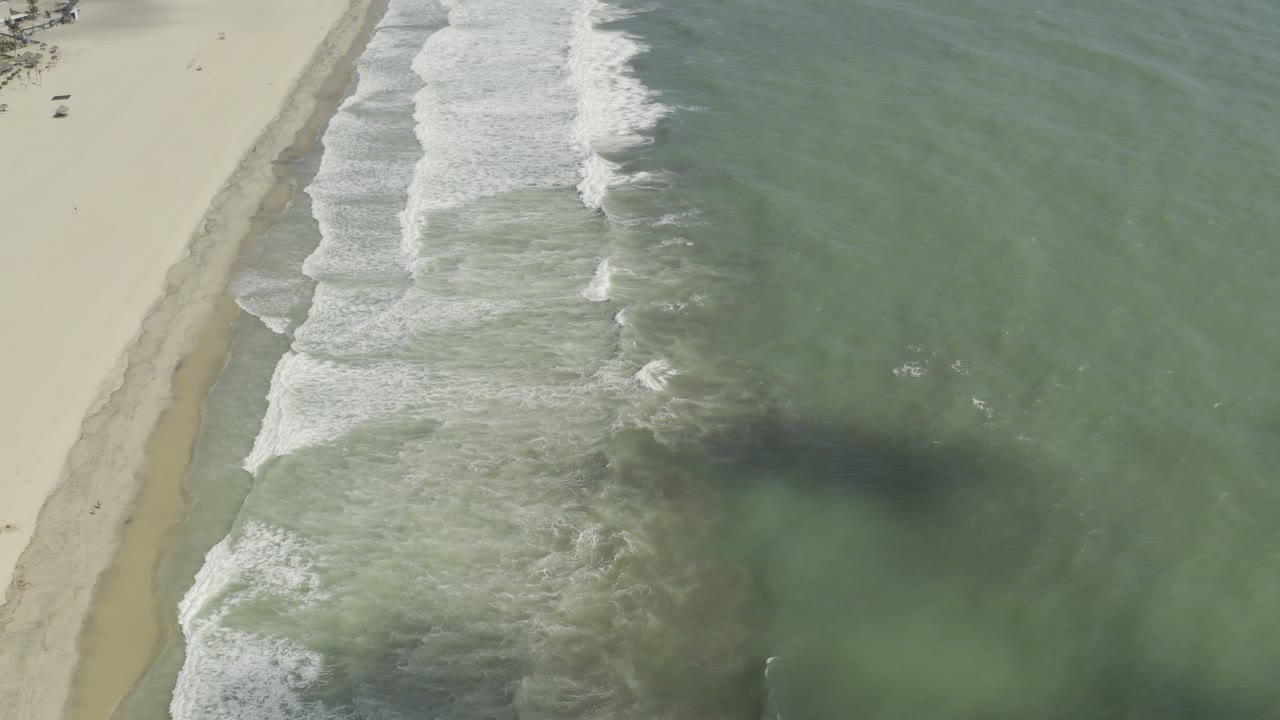 Aerial view over a Los Cabos beach with pale sand on the left, diagonal white surf, green-blue ocean on the right, and no visible people.