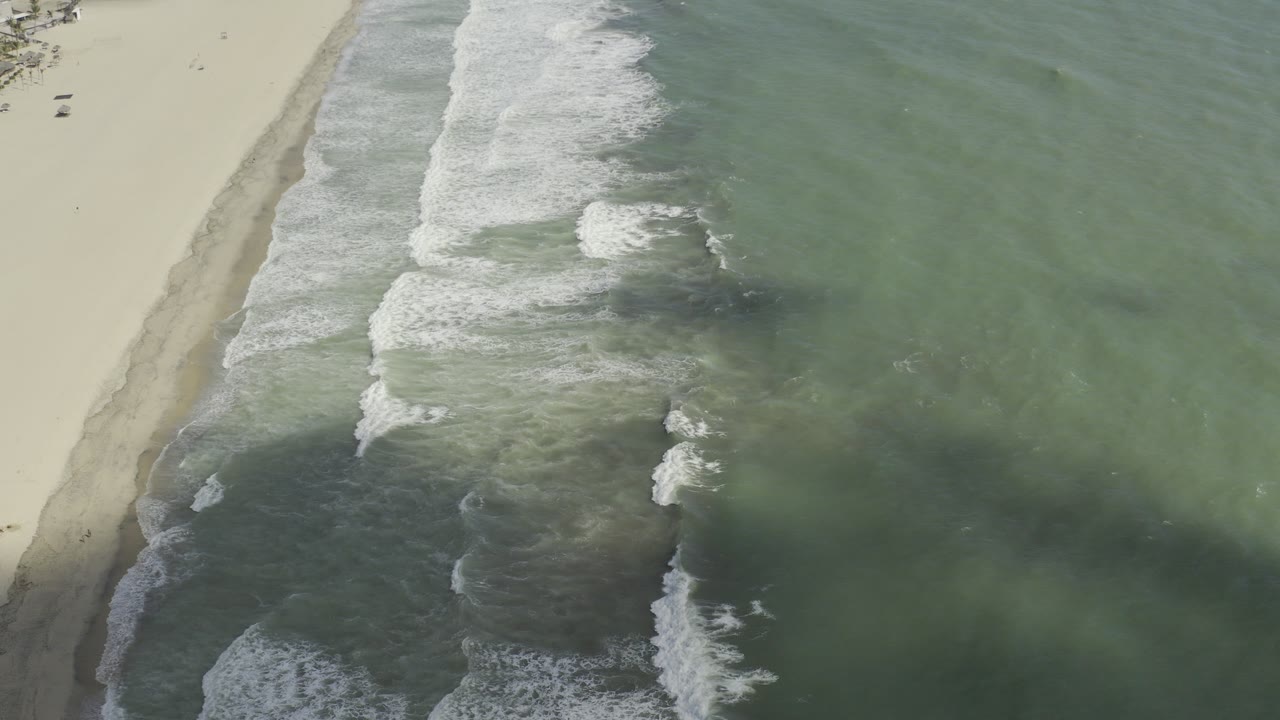 High overhead view of Los Cabos shoreline with pale sand at left, grey-green water at right, and white surf breaking diagonally, with no visible people.