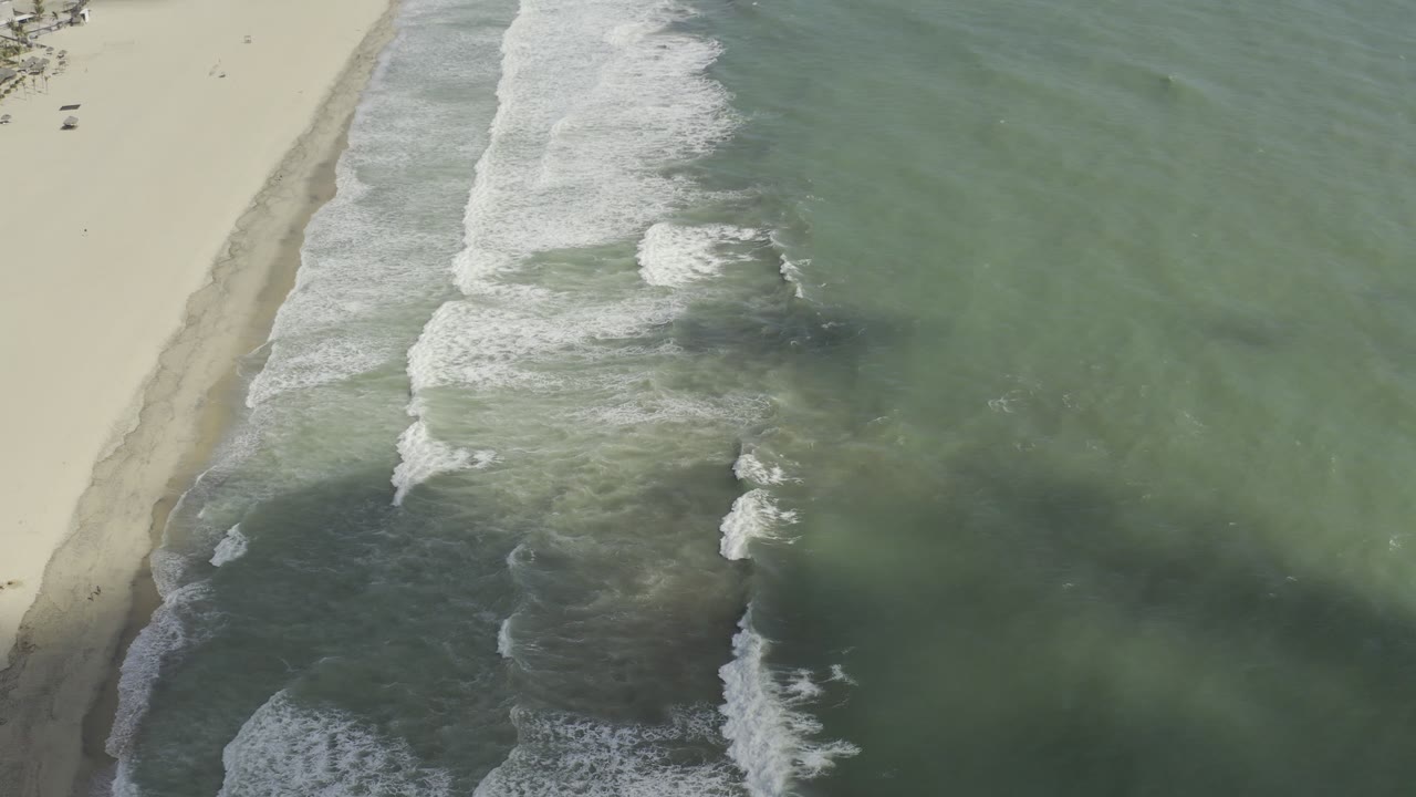 Los Cabos beach seen from above as a broad white wave band rolls toward the sand through grey-green water, with no visible people.