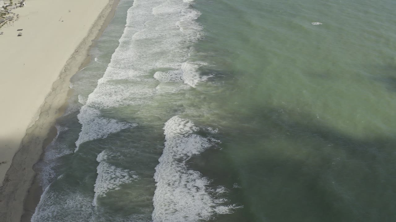 Aerial view of Los Cabos shoreline with pale beach at left, green-blue water at right, white surf crossing the frame, and no visible people.